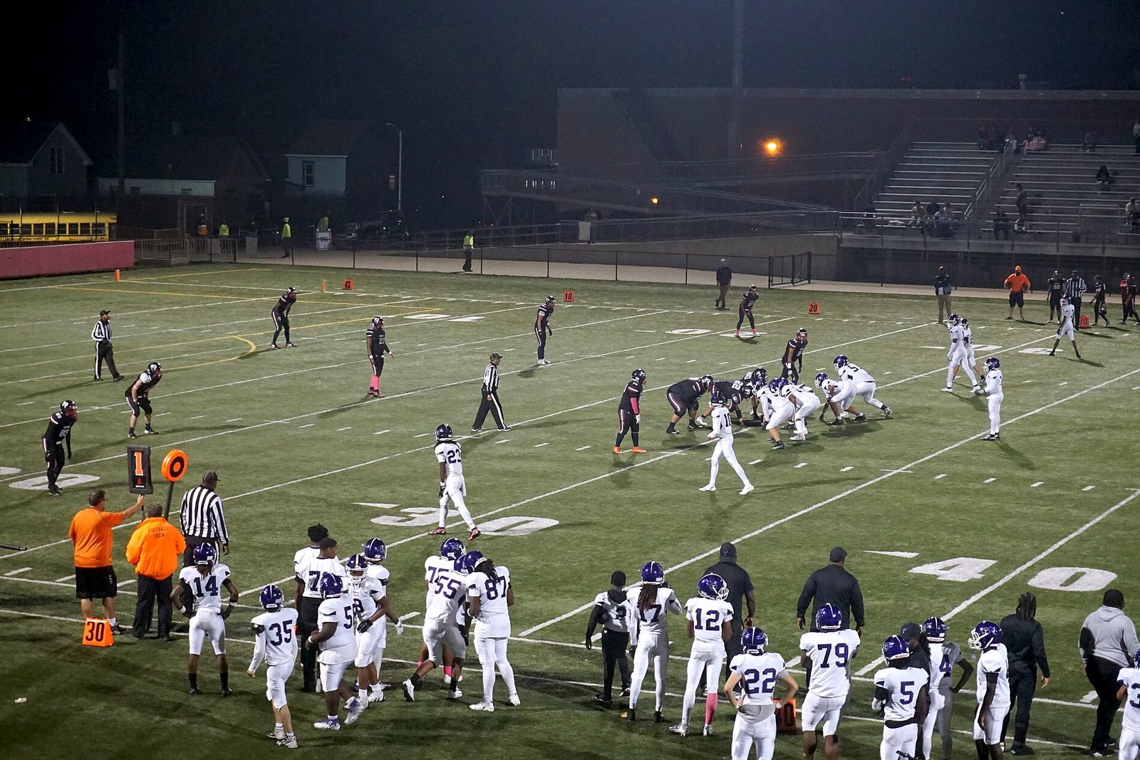 A high school football play on an artificial-turf field at night, with players in white jerseys watching from the sideline.