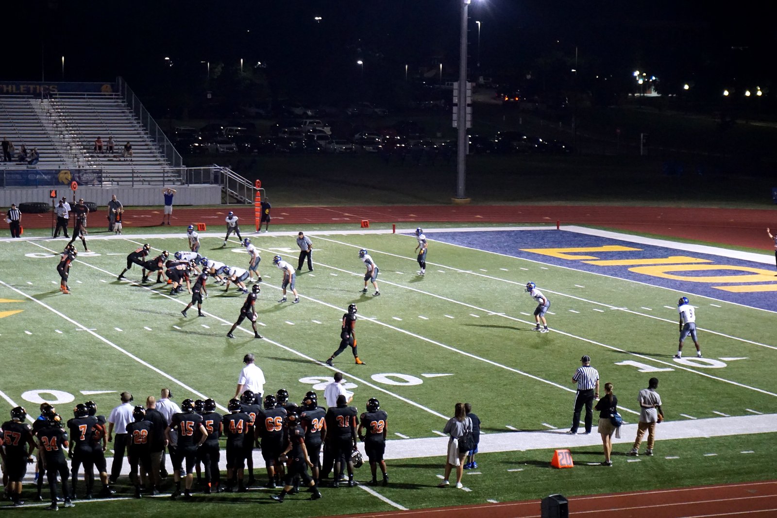 High school football players run a play on a lit field with a painted end zone, as a team in dark jerseys watches from the sideline at night.