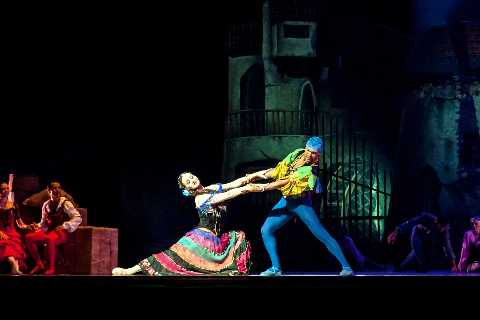Two ballet dancers in colorful costumes perform a duet on a dark stage as other costumed dancers watch from the sides.