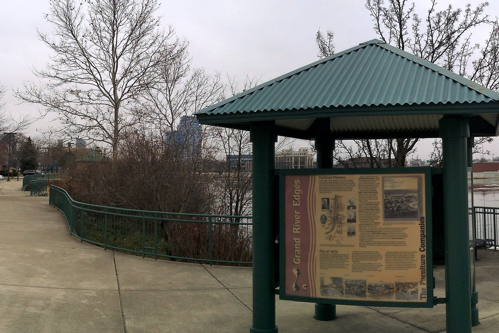 A paved riverside walkway in winter with a green-roofed information kiosk, bare trees and a railing along the Grand River.