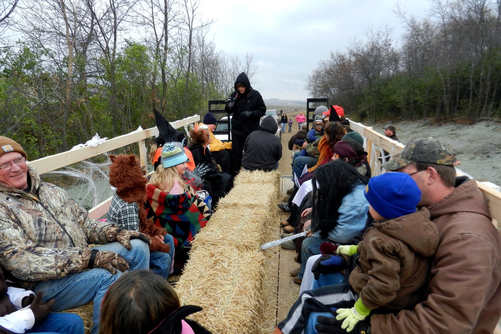 Children in Halloween costumes and adults sit on hay bales on a flatbed trailer decorated with spiderwebs during an outdoor haunted hayride.