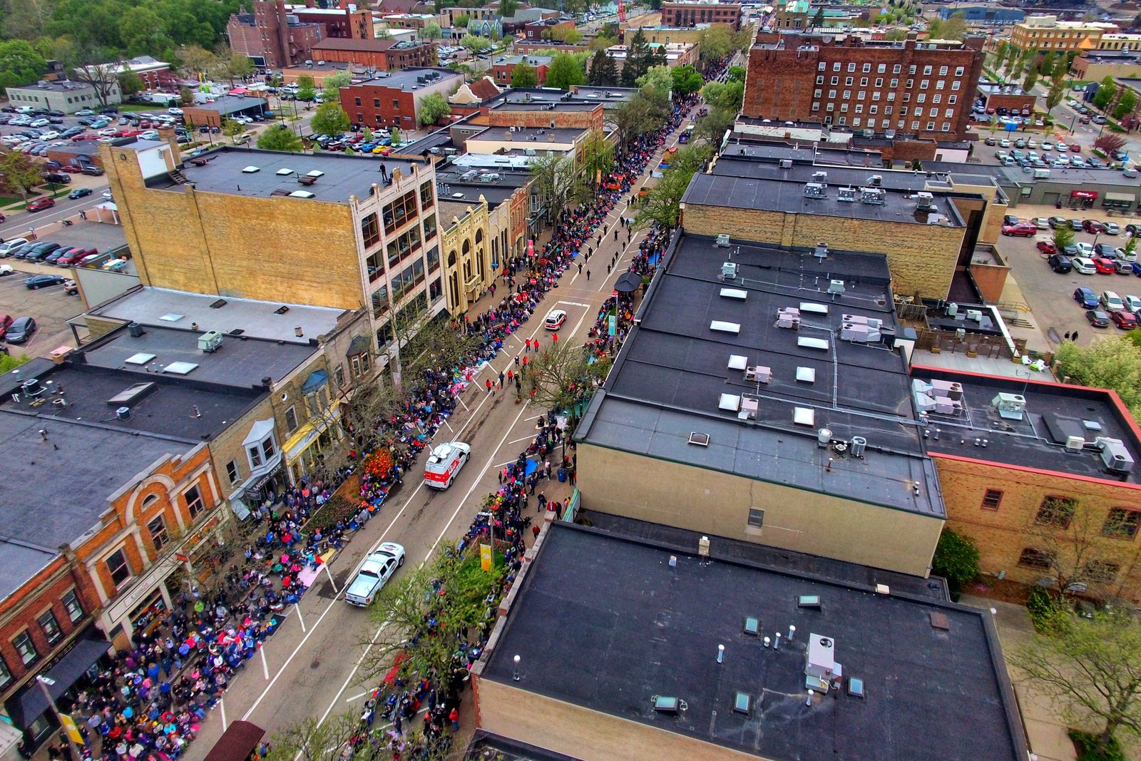 An aerial view of a busy downtown main street lined with brick buildings, with crowds on the sidewalks and trees along the road.