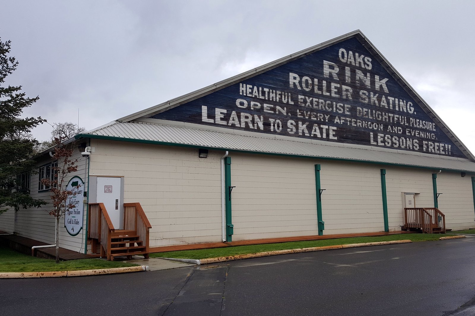 The exterior of a large wood-sided roller rink building with a sign on its peaked roof, behind a parking area and trees.