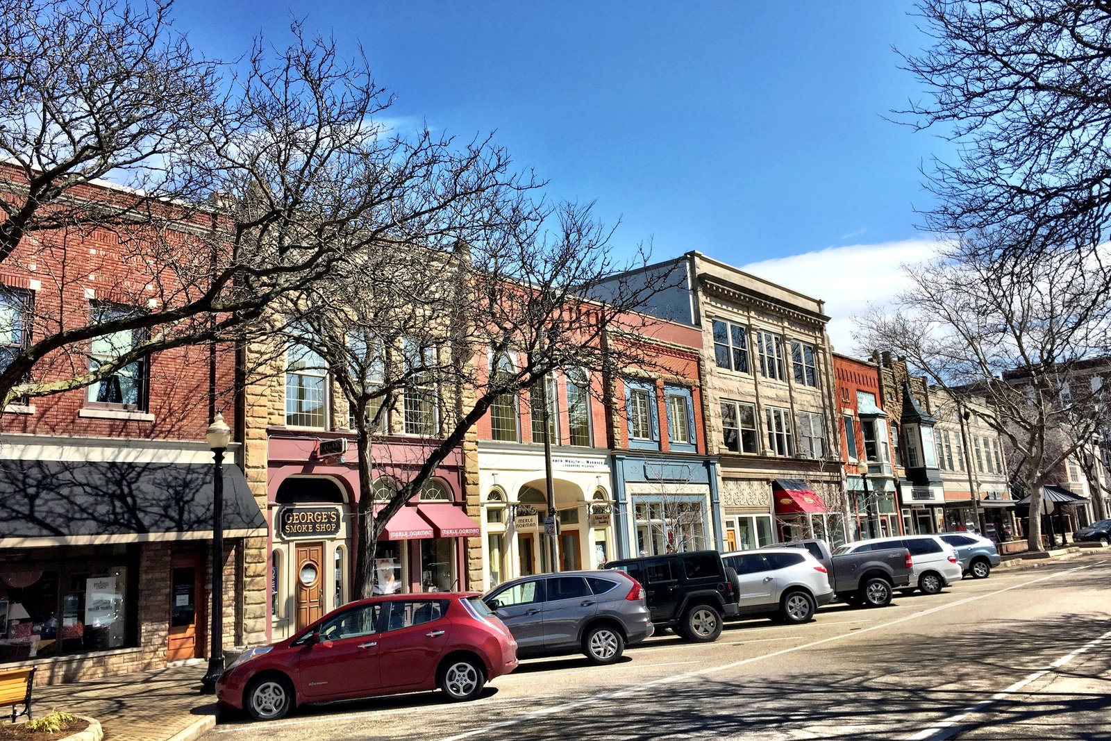 A row of two- and three-story brick storefronts along a downtown street with parked cars and bare trees under a blue sky.