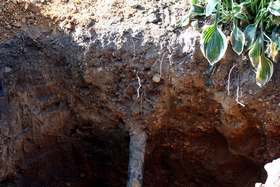 An excavated hole in a city lawn exposing an old gray metal water pipe running toward a house.