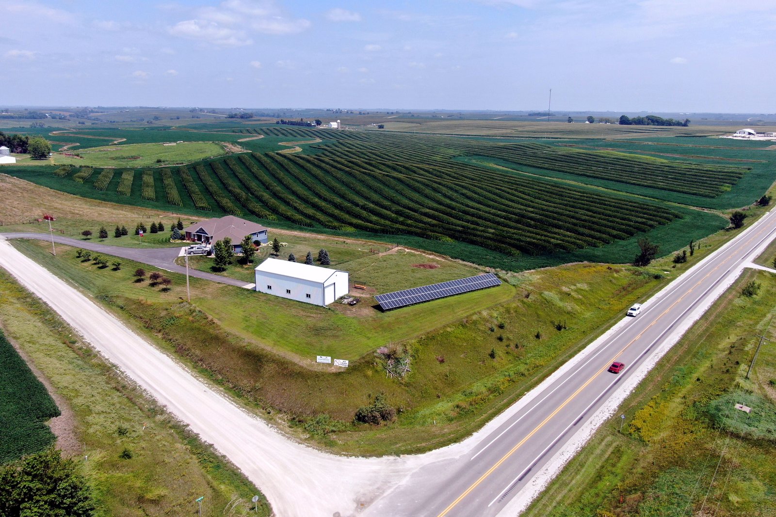 An aerial view of rows of solar panels next to green farm fields, a farm building and a two-lane road.