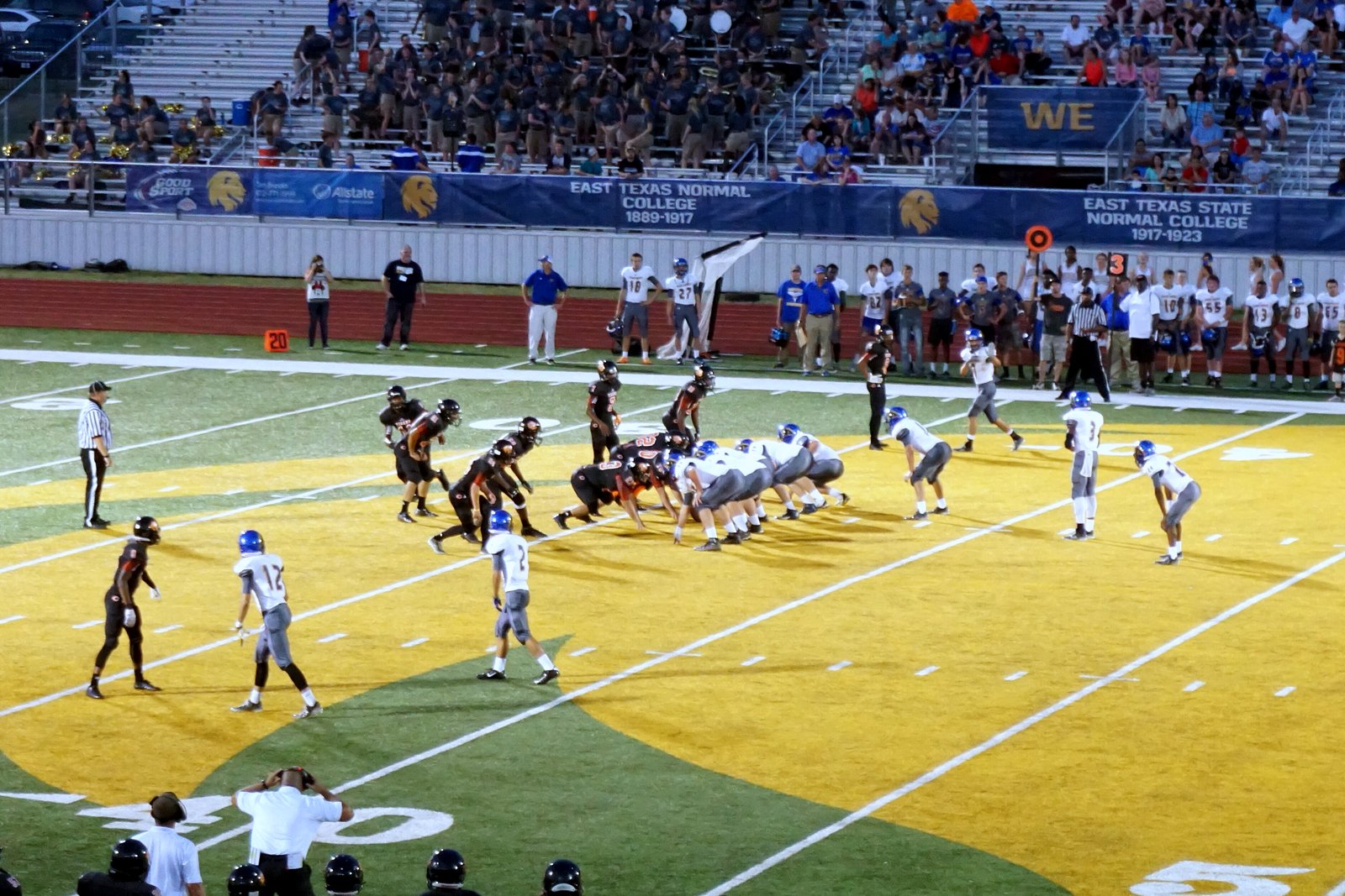 High school football players in white and dark uniforms line up at the line of scrimmage on a green field under stadium lights.