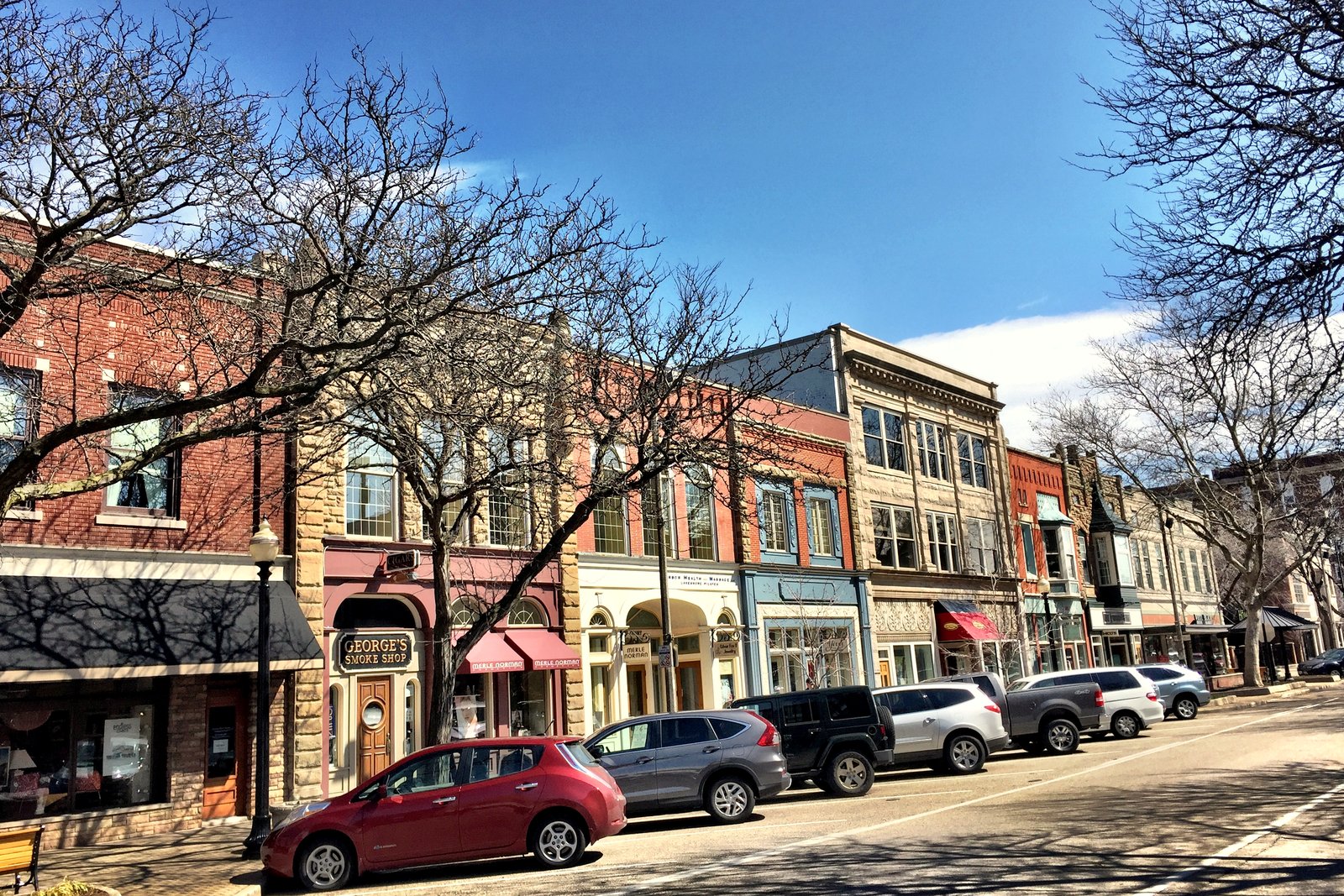 A sunny downtown street lined with two- and three-story brick storefronts, bare trees and cars parked at the curb.