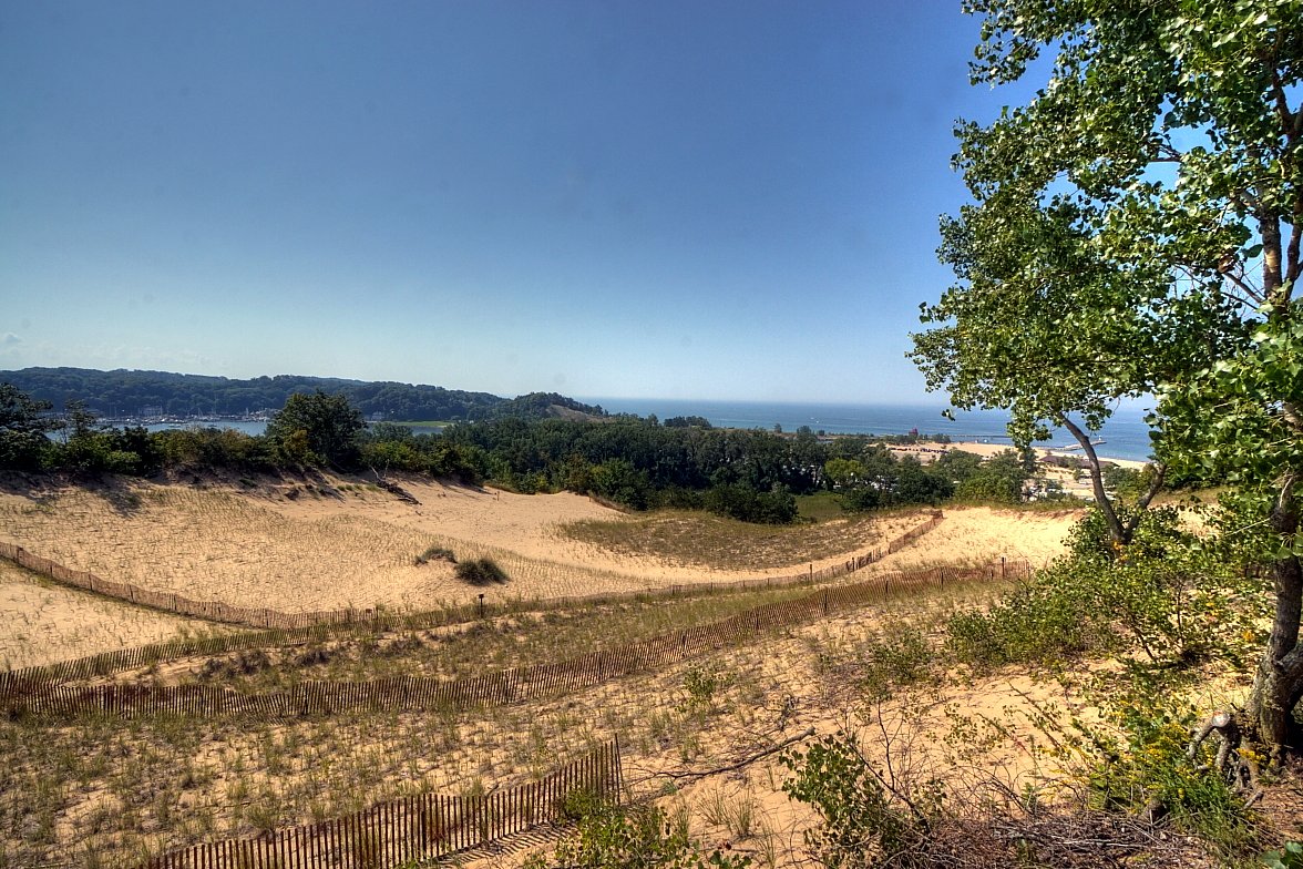 A view from a high sand dune over grass, shrubs and trees toward a lake and a distant shoreline under a blue sky.