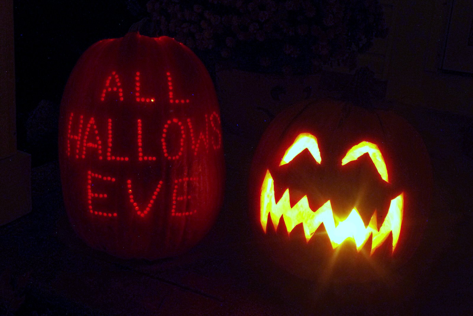 Two glowing carved pumpkins in the dark, one carved with the words All Hallows Eve and the other with a grinning face.