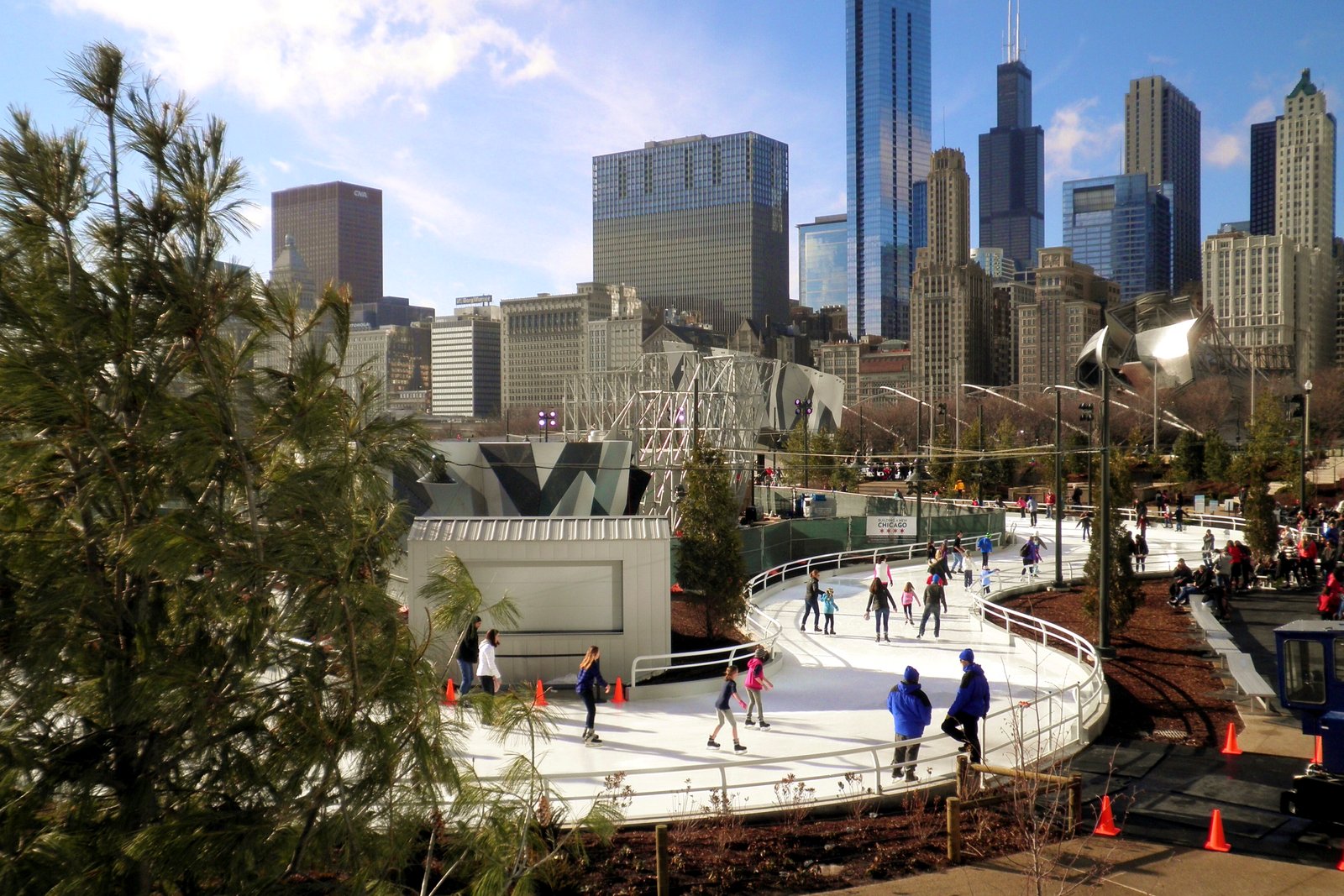 Skaters circle a curving outdoor ice ribbon lined with railings and orange cones in a city park, with pine trees in front and downtown Chicago skyscrapers behind under a blue sky.