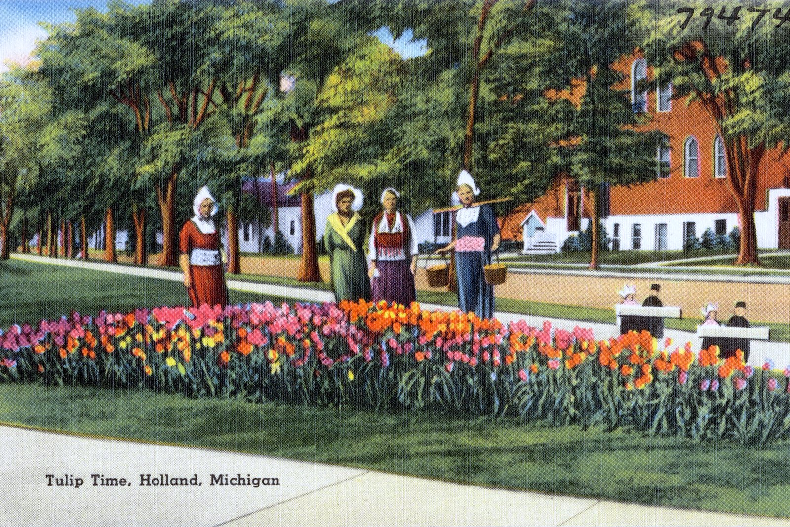 A vintage color postcard of a garden bed of red and yellow tulips in front of trees, with women in traditional Dutch costume standing behind the flowers.