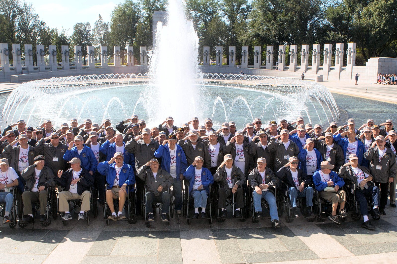 A large group of veterans in blue caps and shirts gathered in front of the fountain at the World War II Memorial in Washington.