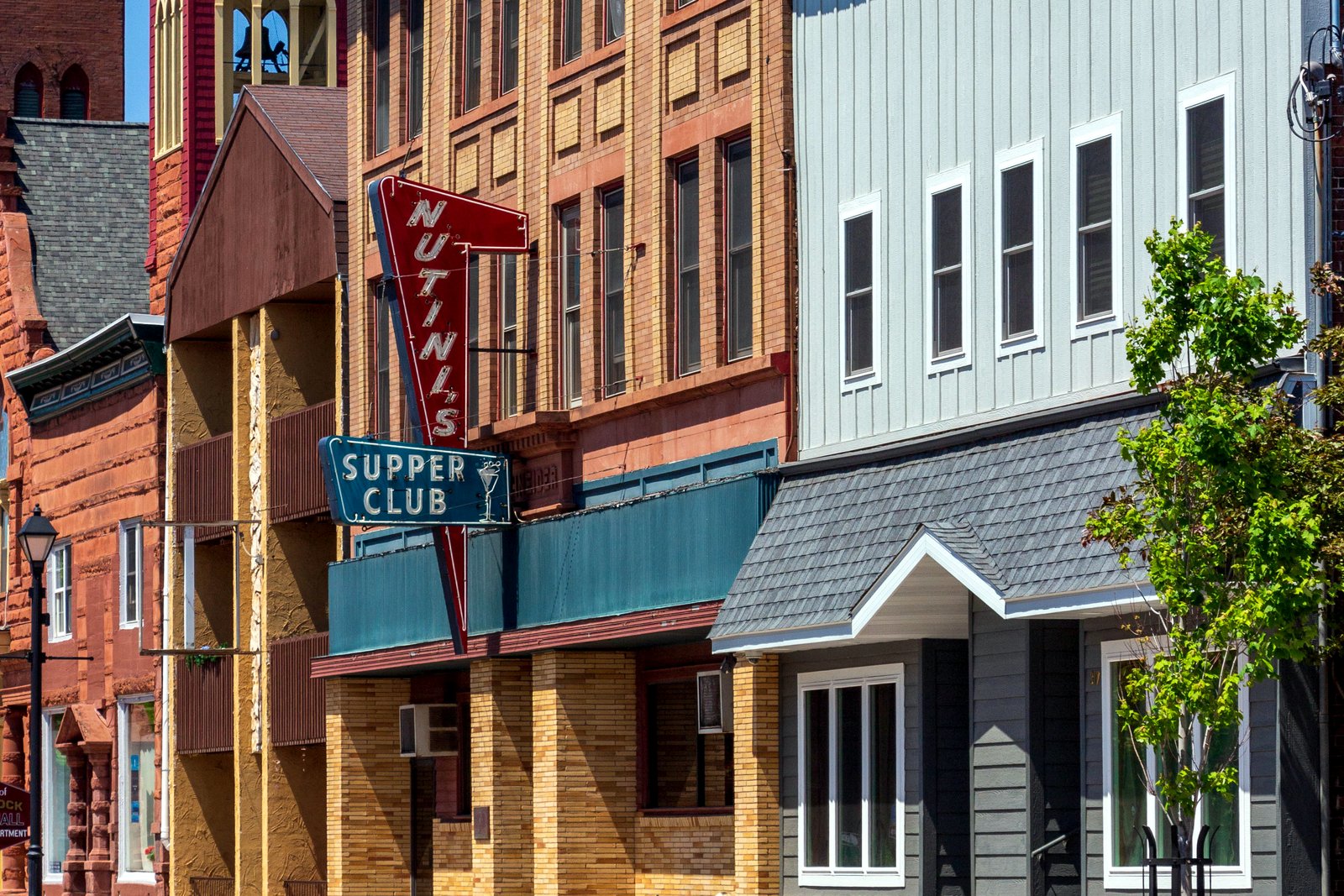 Brick and painted storefront buildings with signs and awnings line a downtown street in a small northern town under a blue sky.