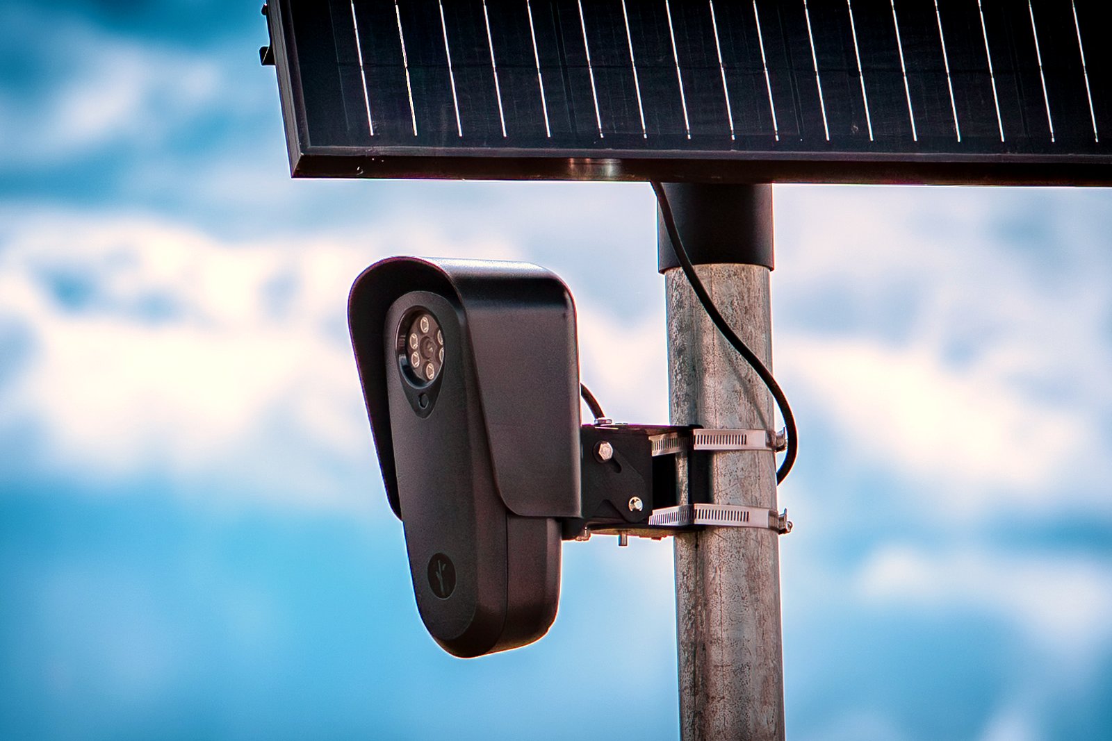 A black Flock Safety license plate reader camera with a small solar panel mounted on a pole against a blue sky.