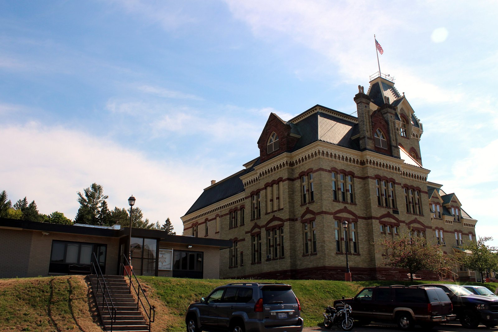 A sandstone courthouse with a tall clock tower beside a plainer brick building that houses the county jail, under a blue sky.
