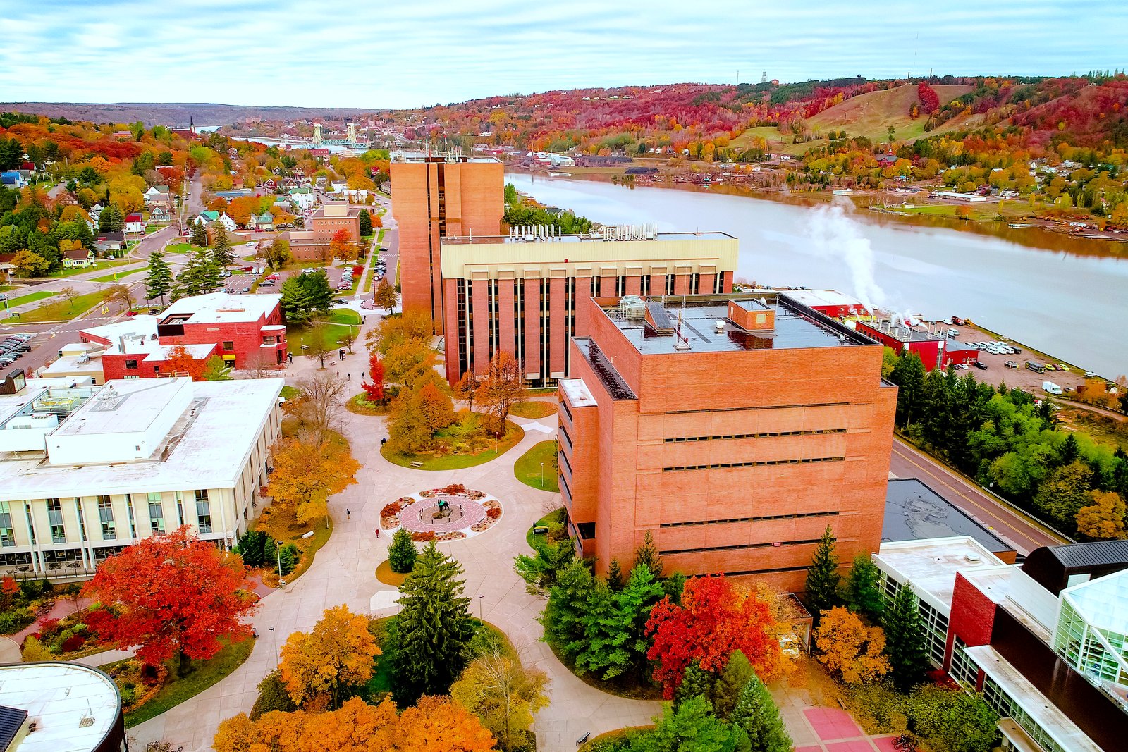 Aerial view of a university campus of brick buildings beside a wide canal, with autumn-colored trees and hills.