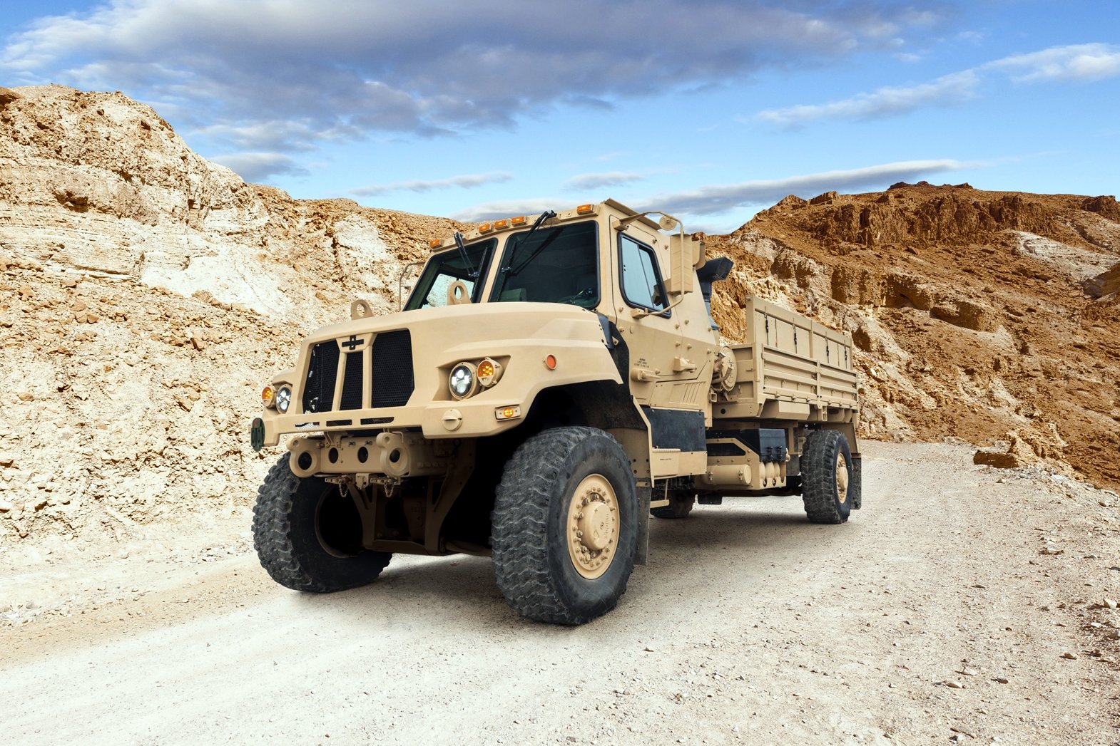 A tan, square-nosed military cargo truck with a canvas-covered bed drives along a dirt road between sandy hills.
