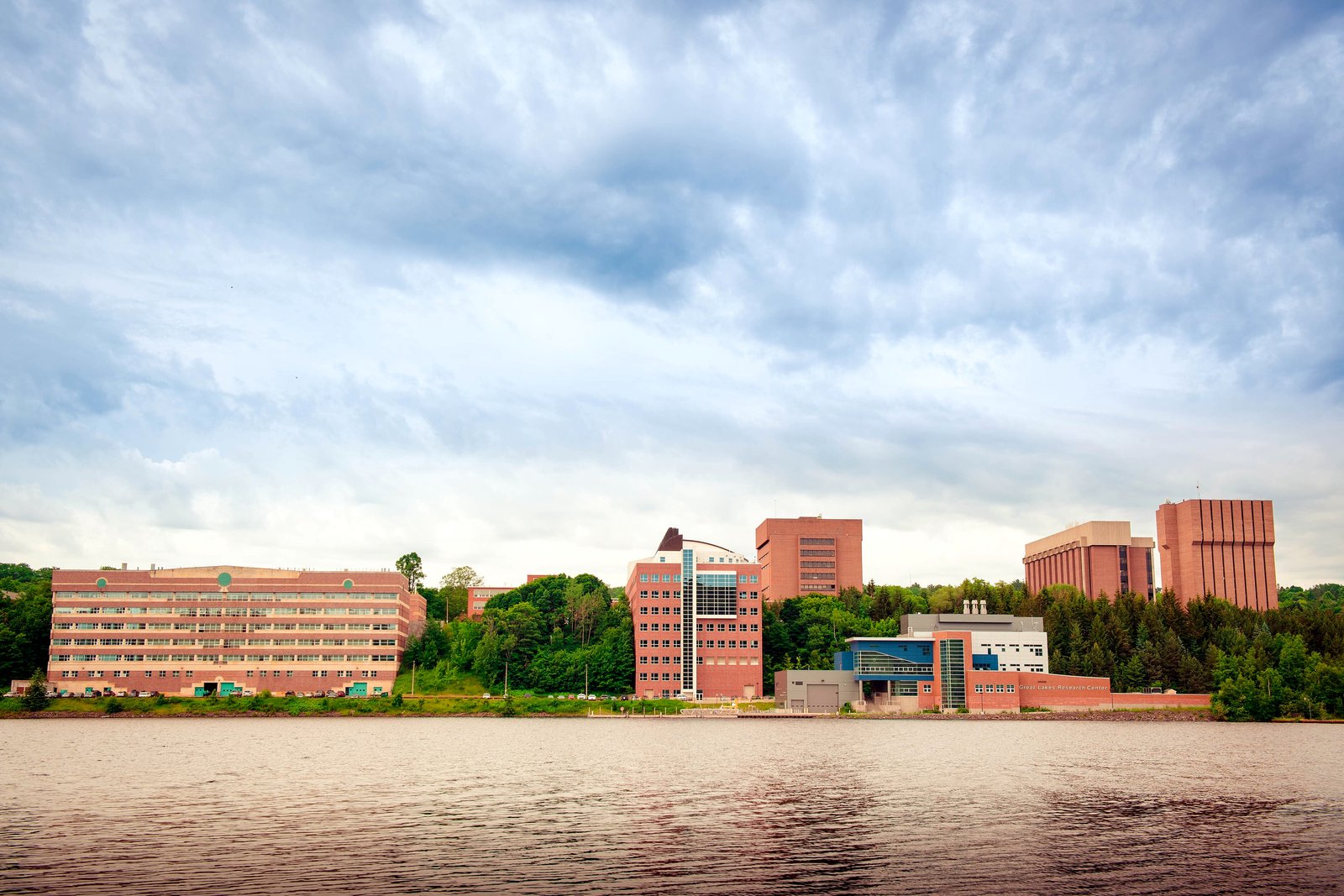 A row of brick and glass university buildings along the water under a cloudy sky, seen from across a canal.