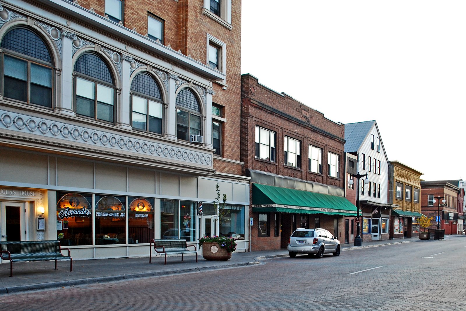 A downtown street of older brick and stone commercial buildings with parked cars along the curb.