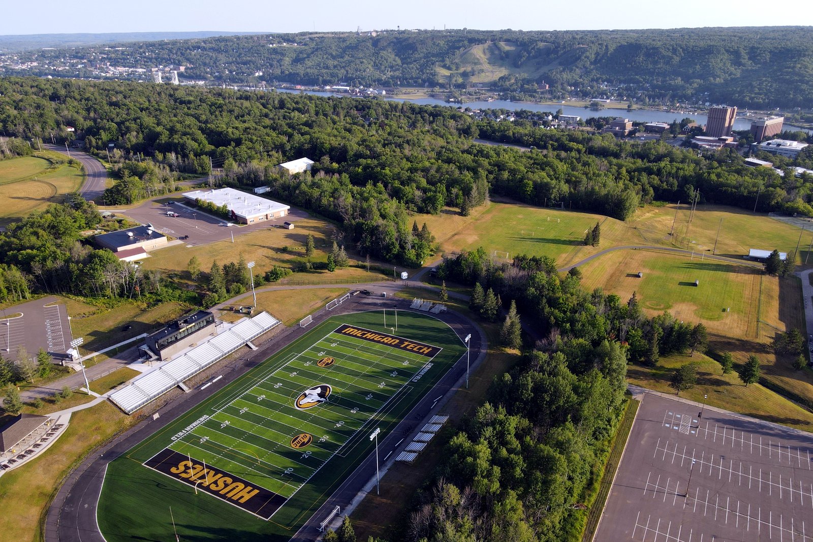 An aerial view of a green football field with a running track, bleachers and a parking lot, surrounded by trees and campus buildings.