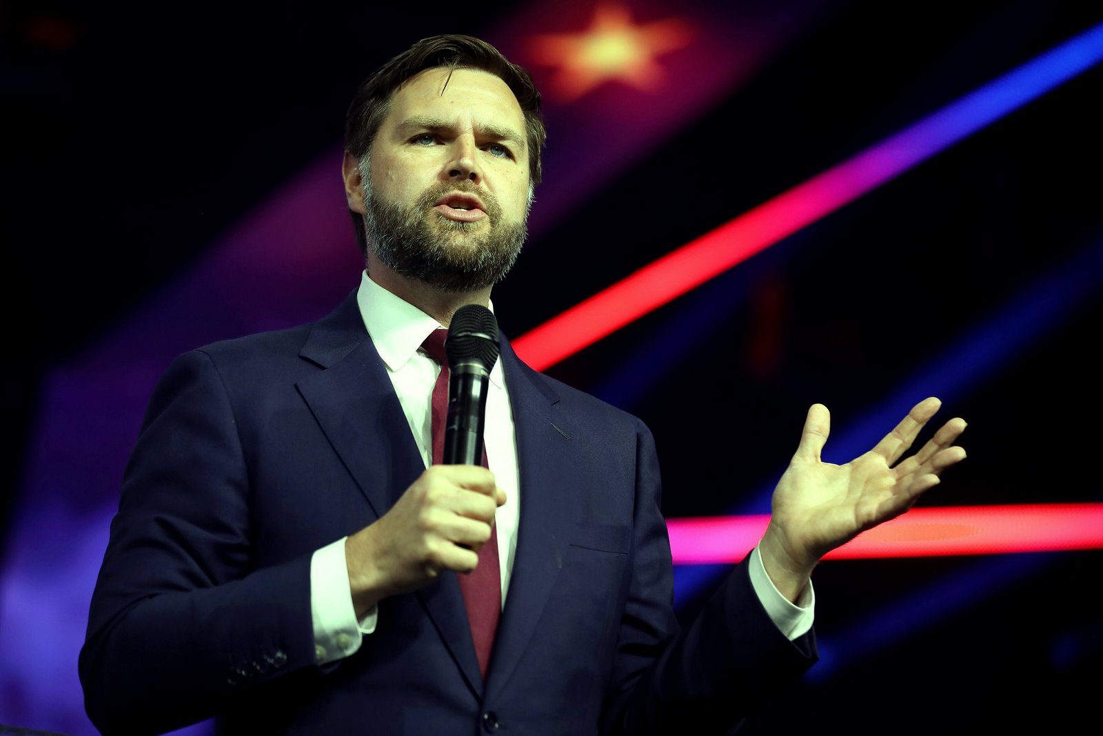 JD Vance, in a navy suit and dark red tie, speaks into a handheld microphone and gestures with his left hand in front of a dark stage backdrop with red and blue light streaks.