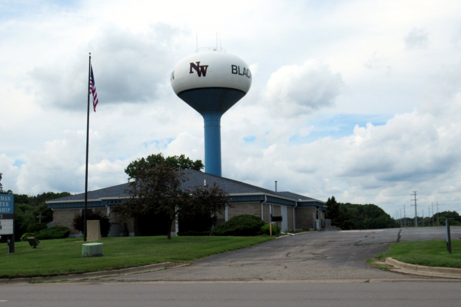 A white water tower on a blue column rises behind the low, gray-roofed offices of Blackman Charter Township, with a flagpole, a driveway and a green lawn in front.