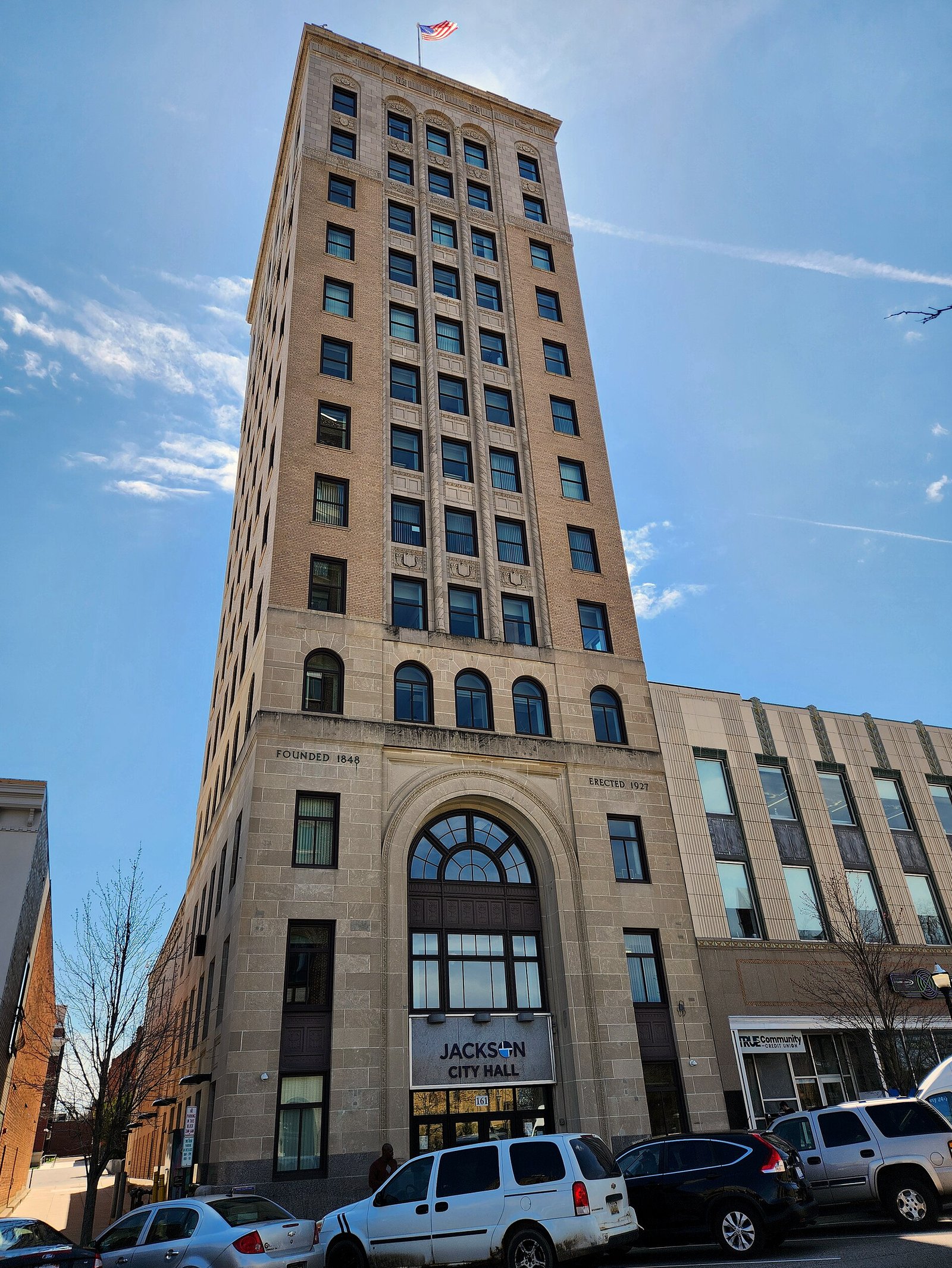 A tall tan brick tower with arched windows and a flag on top, its ground-floor entrance marked Jackson City Hall, with cars parked along the street in front under a blue sky.