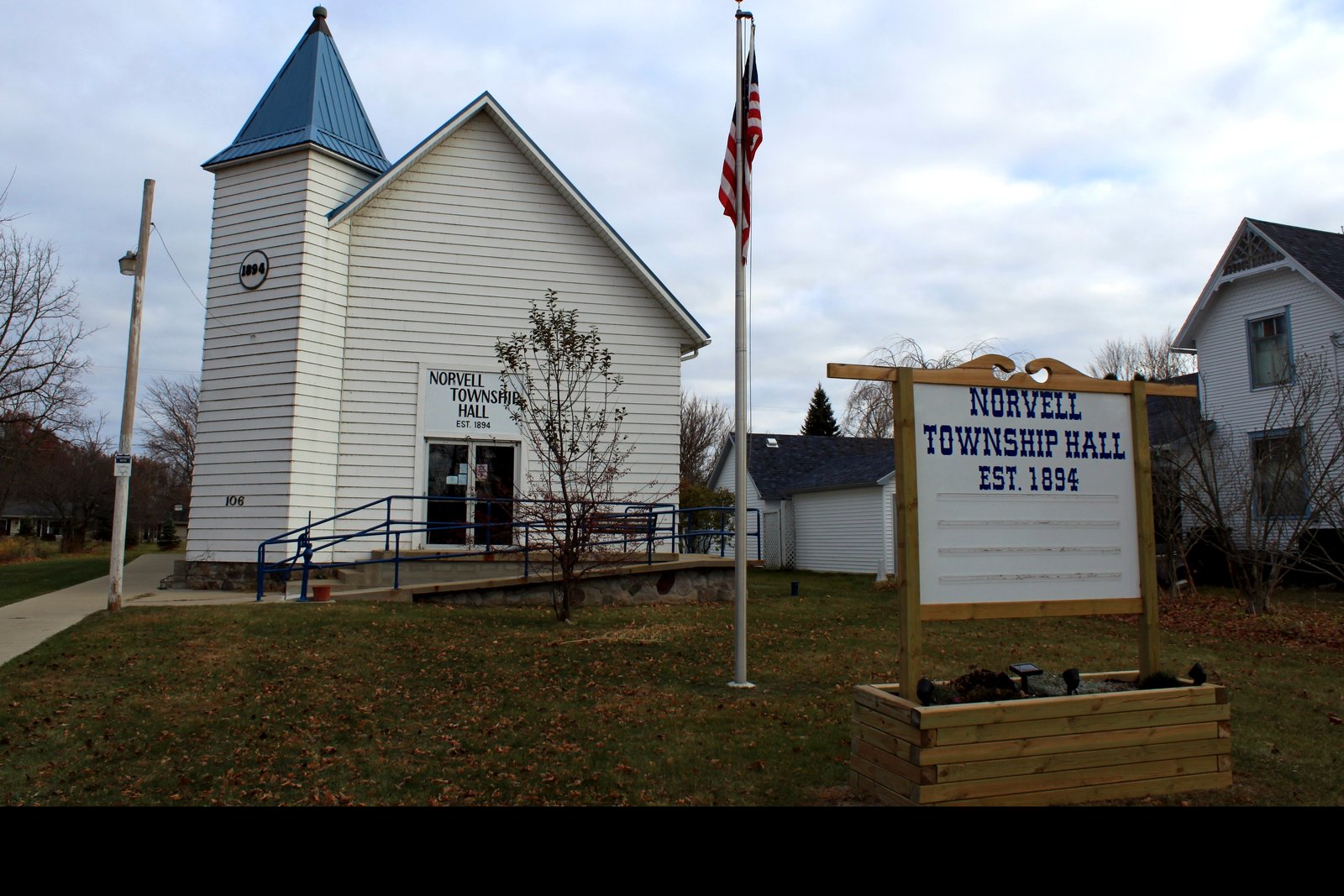 A small white clapboard township hall with a blue-roofed bell tower and a ramp to the front door, an American flag on a pole and a wooden sign reading Norvell Township Hall, Est. 1894.