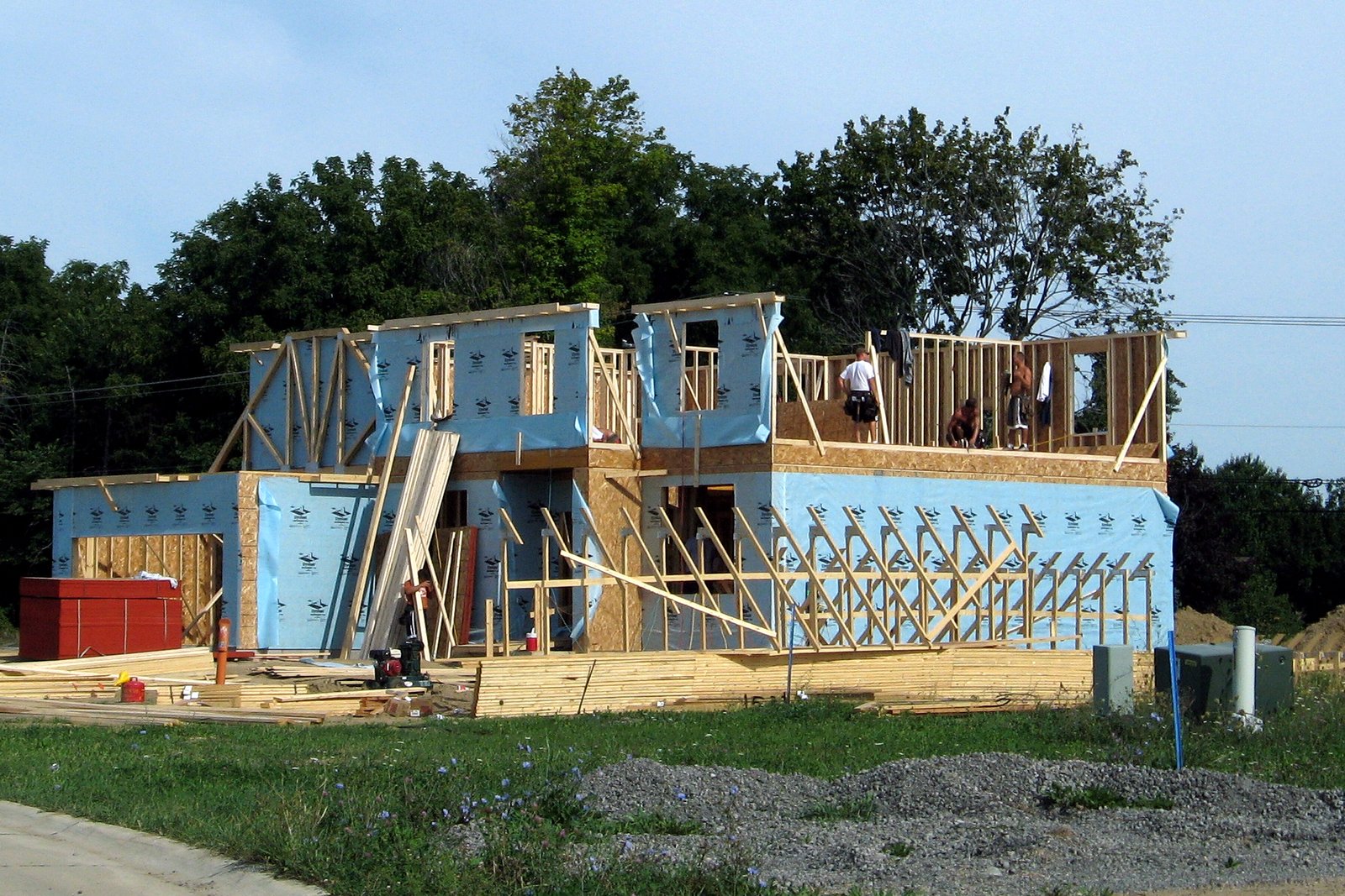 A two-story house under construction, its framing wrapped in blue sheathing, with workers on the upper floor, lumber stacked in front and trees behind.