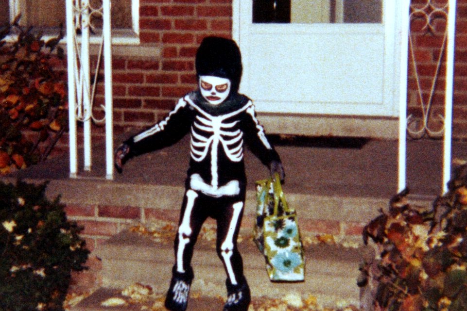 A child in a skeleton costume holds an orange plastic pumpkin bucket while trick-or-treating at night.