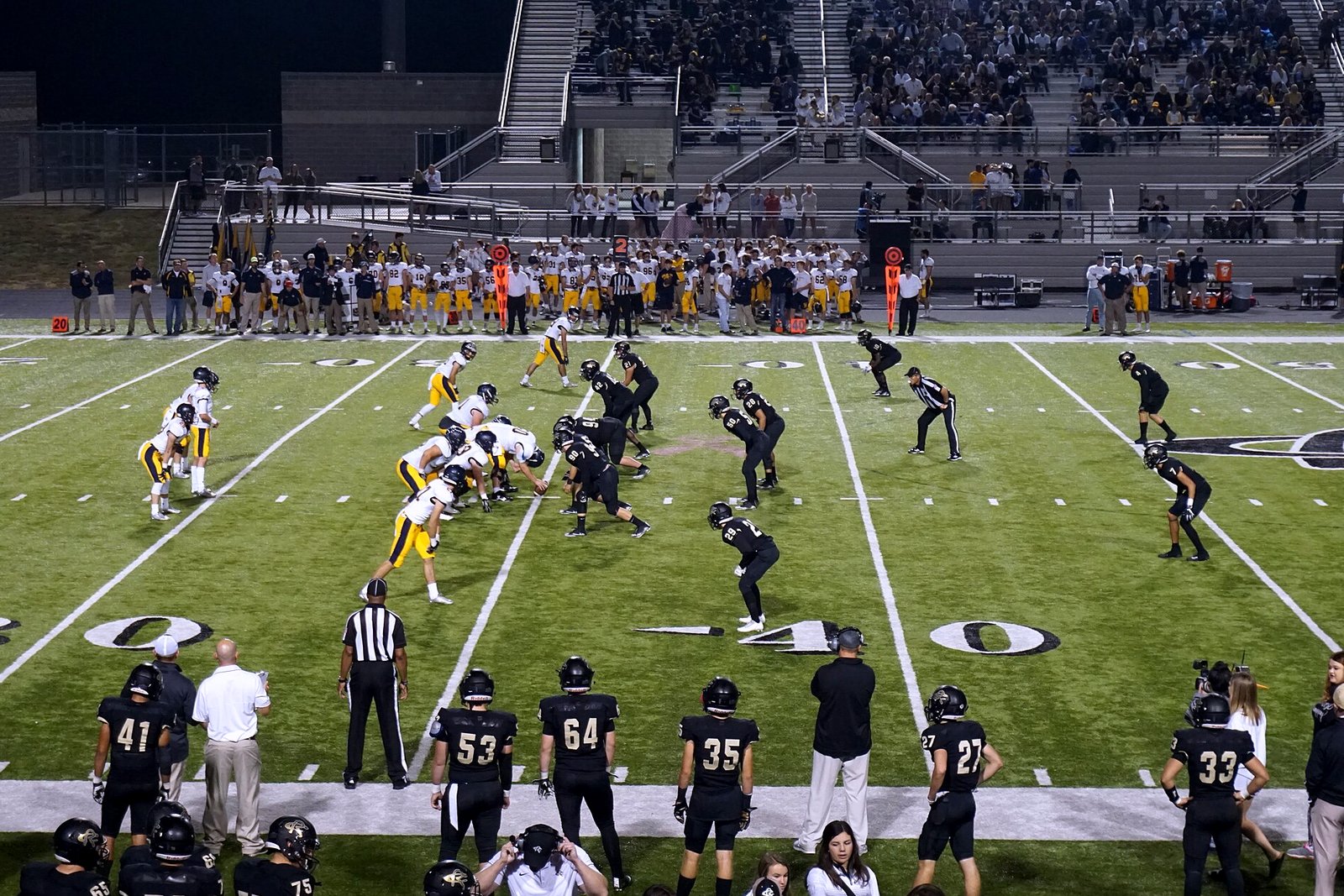 Football players in white jerseys and gold pants line up against a team in black at the line of scrimmage under stadium lights, with a crowded grandstand behind them.