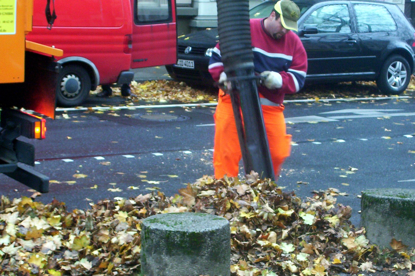A worker in orange pants and a red sweater guides a large black vacuum hose over a pile of fallen leaves at the edge of a street, with a truck and parked cars behind him.
