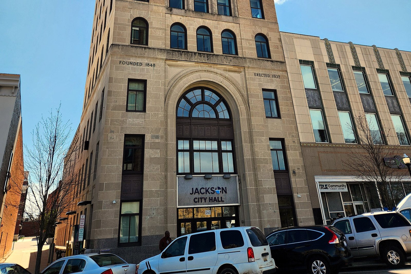 Jackson City Hall’s stone entrance with a “Jackson City Hall” sign over the doors and cars parked along the curb.