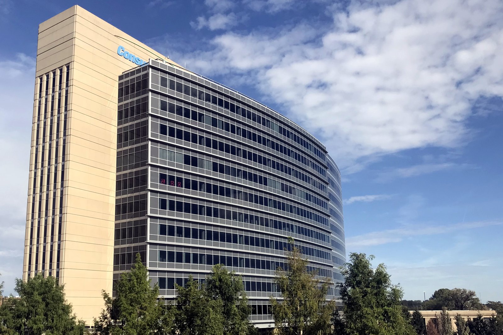 A tall cream-colored office tower with a curved glass building beside it and a Consumers Energy sign near the top, behind a row of young trees under a blue sky.