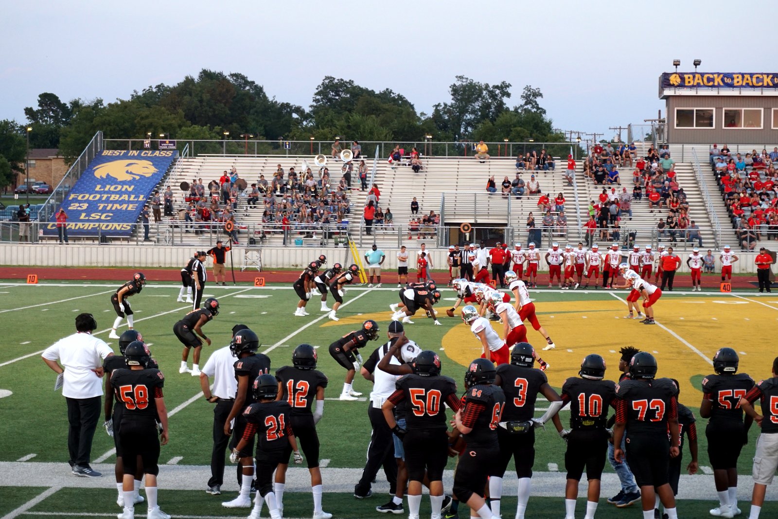 High school football players line up at the line of scrimmage under early evening light, with a team in dark jerseys watching from the sideline, in a file photo.