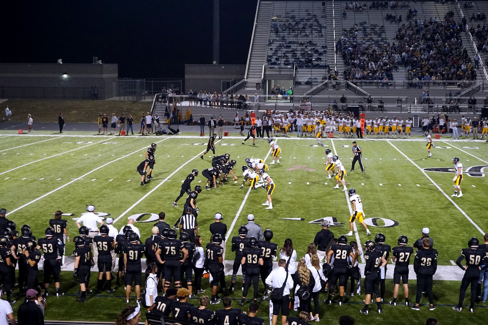 A high school football team lines up on offense on a lit turf field as a team in black watches from the sideline in the foreground.