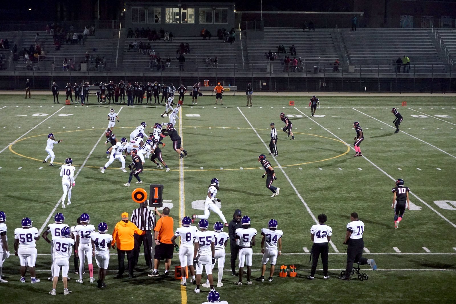 High school football players run a play on a turf field under stadium lights, with a team in white jerseys along the sideline in front.