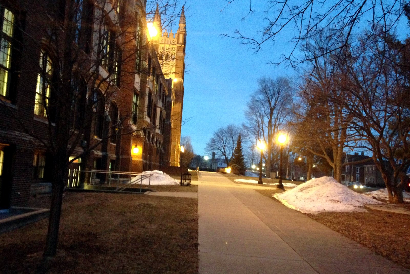 A long brick school building with Gothic-style towers and lit lamps along a sidewalk at dawn, with patches of snow on the lawn, at Jackson High School.