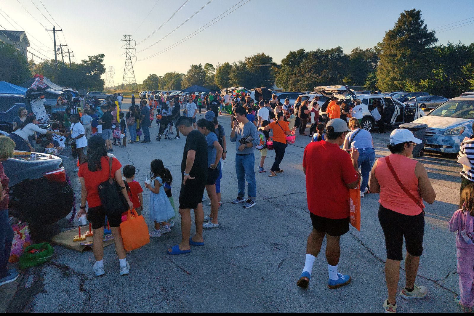 Families and children in costumes crowd a parking lot lined with decorated car trunks at a trunk-or-treat event in the early evening.