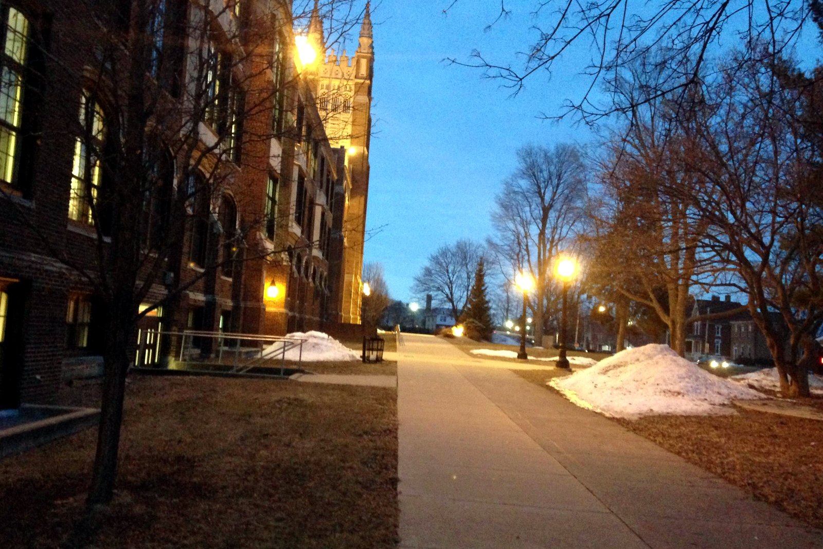 A brick school building with a stone Gothic tower, lit lamps along a long sidewalk and piles of snow on the lawn in dim early light.