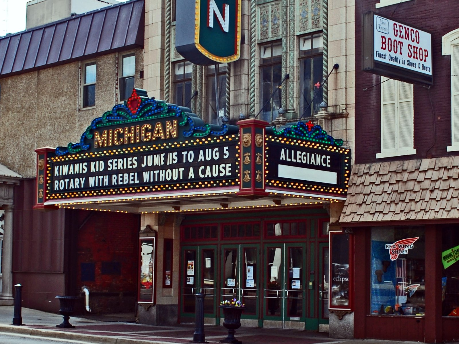The ornate cream-colored terra-cotta front of the Michigan Theatre in downtown Jackson, with a lighted marquee spelling Michigan in gold letters above green-framed glass doors, a boot shop with a Red Wing sign on the right and an empty sidewalk in front.