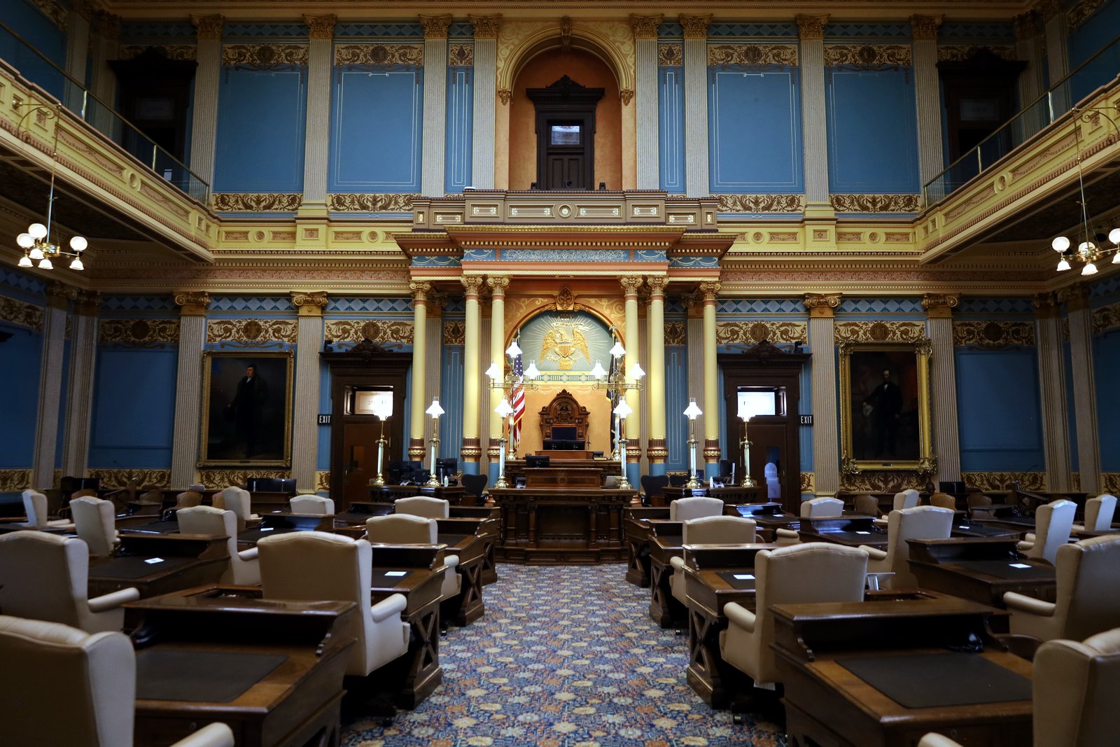 The empty Michigan Senate chamber, with rows of wooden desks and cream leather chairs facing a raised rostrum and an American flag, framed by blue walls, gold columns, portraits and upper galleries.