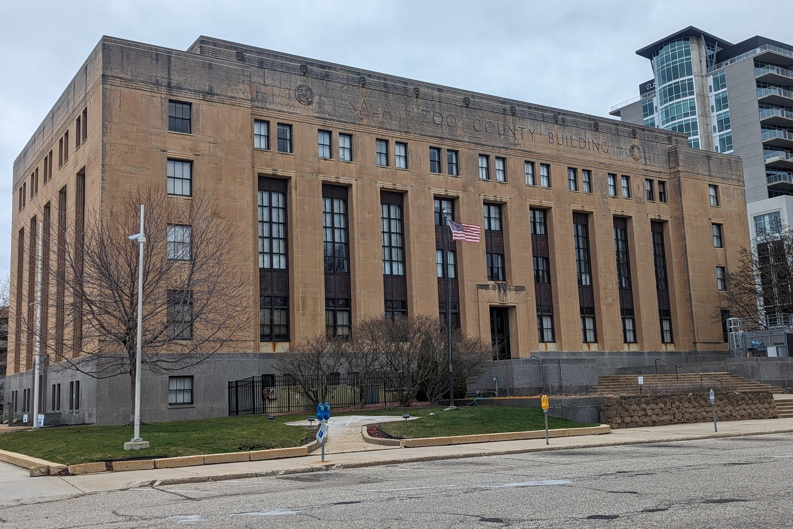 The Kalamazoo County Building, a tan stone Art Deco government building with tall vertical windows and an American flag, on an overcast day in downtown Kalamazoo.