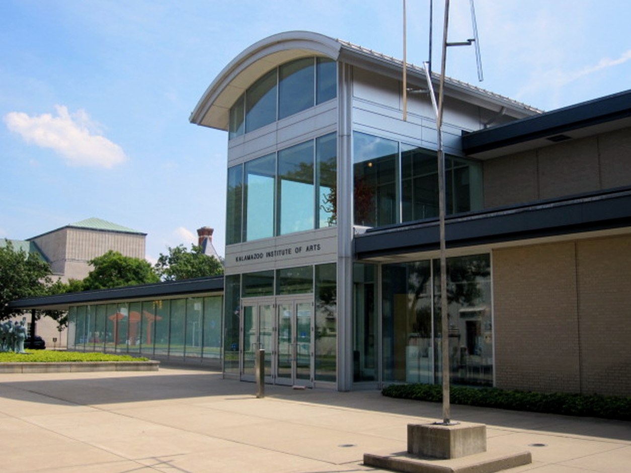 A modern museum entrance with a tall curved glass tower labeled Kalamazoo Institute of Arts, glass walls and a tan brick wing, a wide concrete plaza in front under a blue sky.
