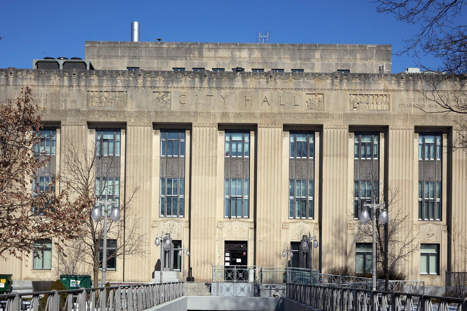A limestone Art Deco building with CITY HALL carved above tall vertical window bays, seen from a walkway with iron railings.