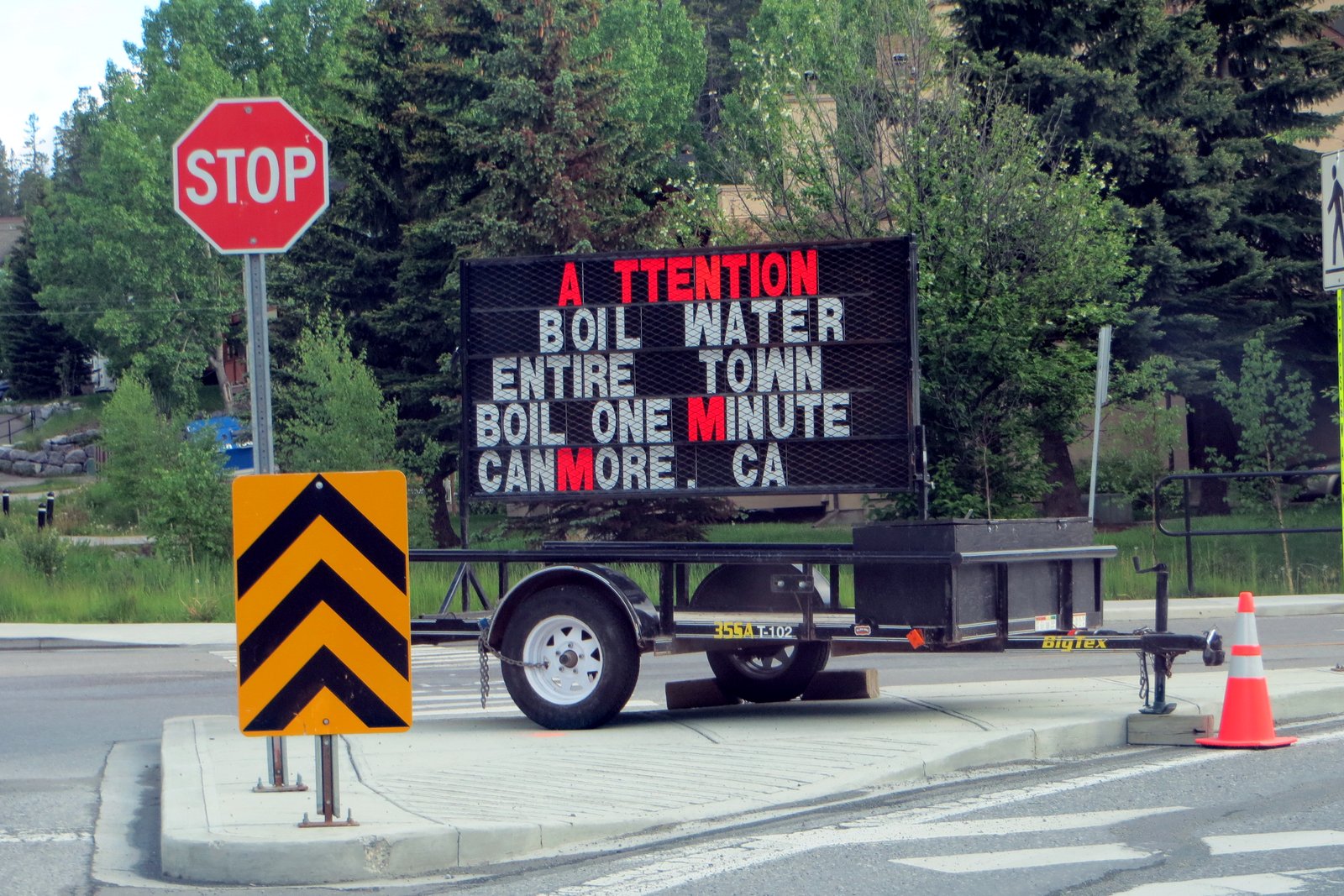A portable electronic road sign on a trailer at a street corner reads Attention, Boil water, entire town, boil one minute, next to a stop sign and an orange traffic cone.