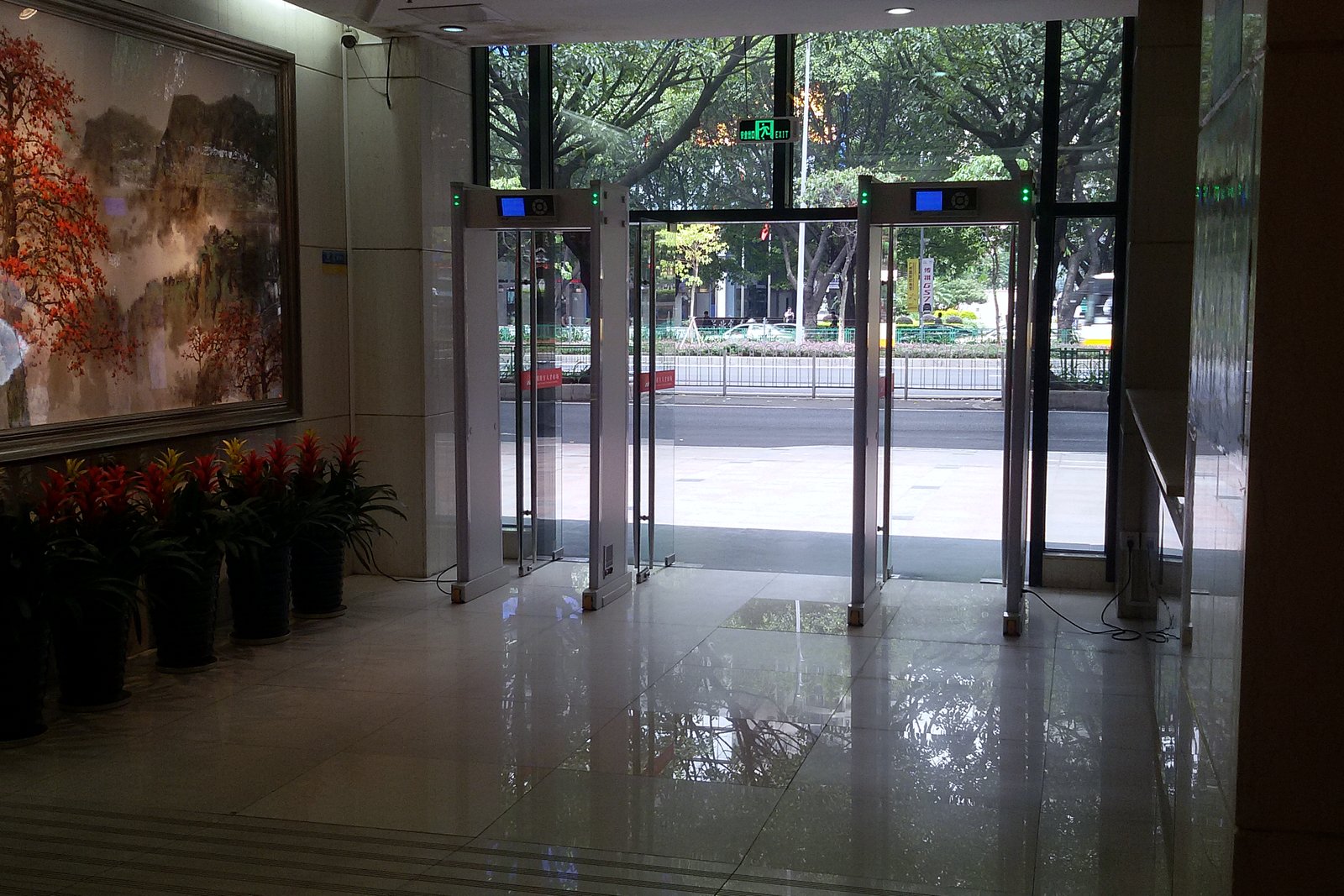 Walk-through metal detector gates inside the glass-walled lobby of an office building, with potted red flowers and a large painting on the wall.