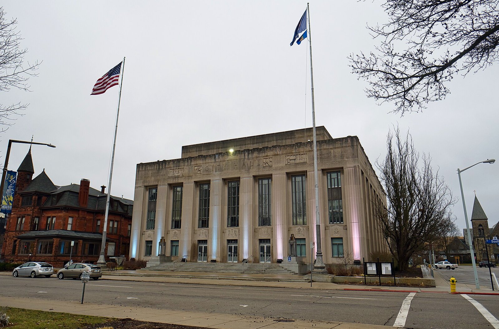 Kalamazoo City Hall, a pale Art Deco stone building with tall vertical windows and two flagpoles, on a gray March day, with a red brick Victorian building beside it.