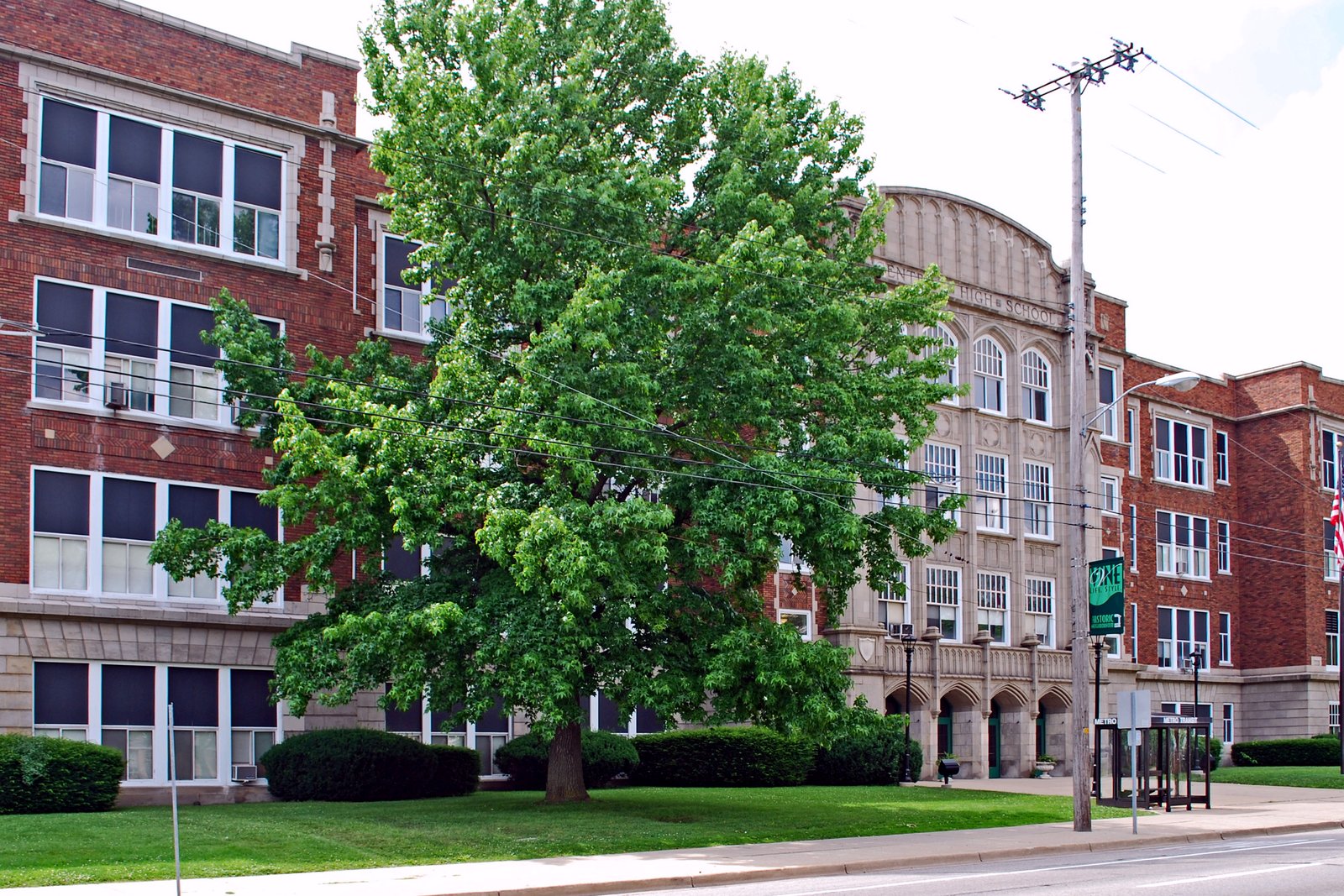 A long three-story red brick and stone school building with tall windows and an arched stone entrance, partly hidden behind a large green tree.